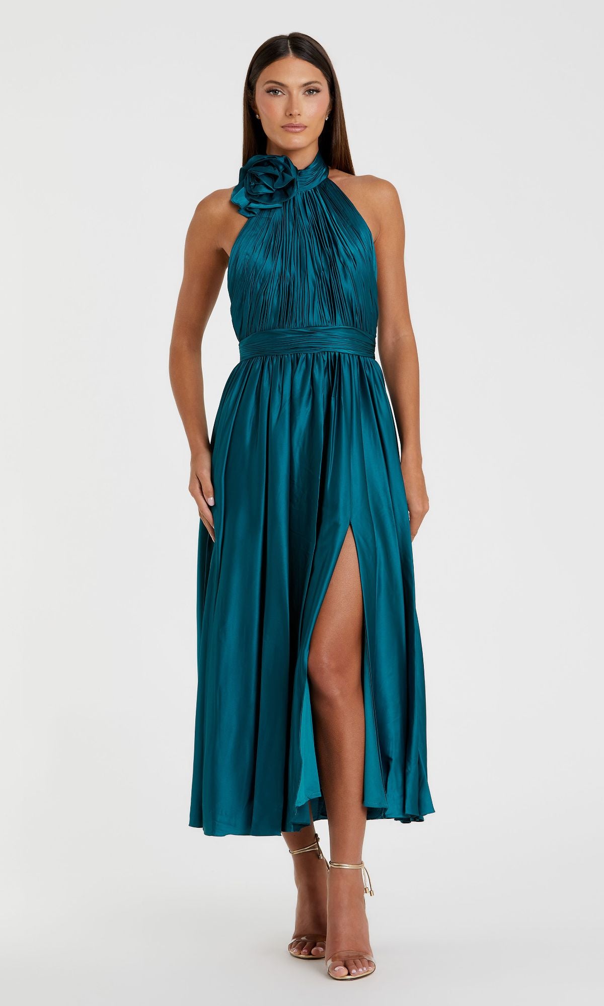 A woman in the Mac Duggal 49674 Halter Midi Wedding Guest Dress—teal, sleeveless with a floral shoulder detail, pleated bodice, and thigh-high slit—creates an elegant semi-formal look paired with strappy heels.